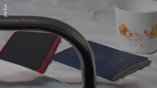 Un passeport ukrainien est posé sur un lit, accompagné d'un téléphone portable et d'une tasse illustrée, dans le cadre du reportage Journal d'un voyage à Marioupol.