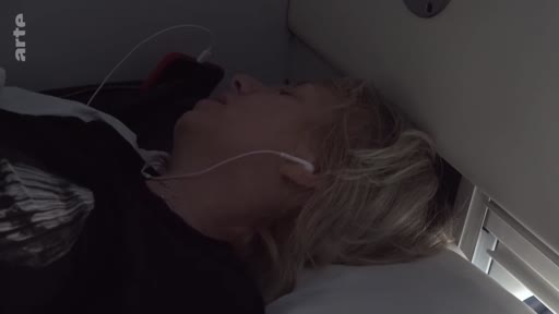 Une femme blonde dort allongée sur un siège de train, portant des écouteurs, lors d'un voyage vers Marioupol. Cette image illustre les conditions de transport et la fatigue durant le périple documenté dans le film Journal of a journey to Mariupol.