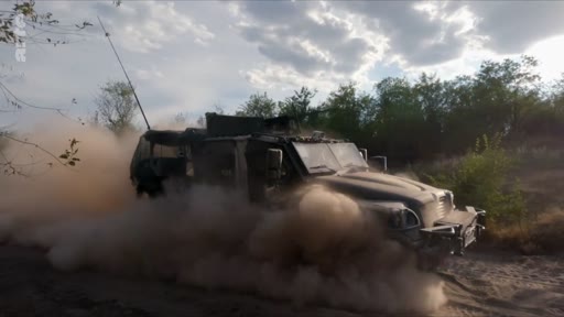 Un véhicule blindé de type Humvee ou similaire circule à vive allure sur un chemin de terre, soulevant un important nuage de poussière dans un environnement rural. Cette image illustre les opérations de transport et de mobilité des troupes sur le terrain lors d'un conflit armé.