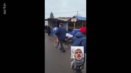 Un homme filme un marché en extérieur, probablement en Ukraine, tout en apparaissant en incrustation vidéo lors d'un appel ou d'un reportage. On aperçoit des étals de marché et des passants dans un contexte de vie quotidienne en temps de guerre.