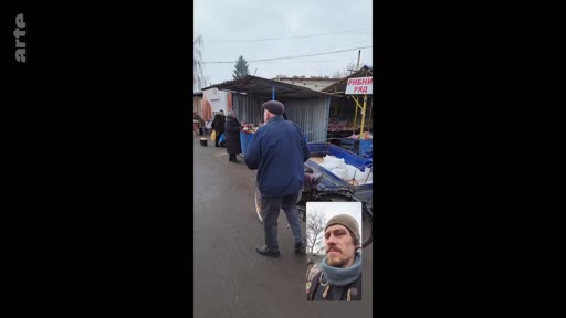 Un homme filme un marché en Ukraine, probablement dans une zone touchée par le conflit, tout en apparaissant en incrustation vidéo (selfie). Le panneau « РИБНИЙ РЯД » (rayon poissonnerie) est visible sur le stand du marché.