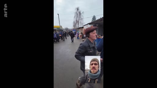 Un homme filme un marché en extérieur tout en se montrant en incrustation vidéo, illustrant le quotidien dans un contexte de crise ou de conflit. La scène montre une interaction entre le terrain et le témoin, typique d'un reportage sur l'adaptation à une nouvelle réalité.