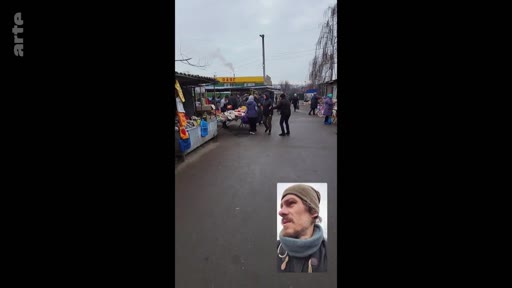 Un homme barbu portant un bonnet et une écharpe filme un marché en extérieur lors d'un appel vidéo, illustrant la vie quotidienne dans un contexte de crise ou de conflit. La scène montre des étals de marché et des passants dans une rue, capturée via une interface d'appel vidéo.