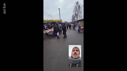 Un homme barbu avec un tour de cou filme un marché en extérieur lors d'un appel vidéo, montrant des étals et des passants dans une ambiance quotidienne.