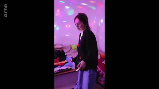 Une jeune femme se tient debout dans une pièce à l'ambiance festive, éclairée par des projecteurs colorés, évoquant un moment de vie communautaire ou de détente.