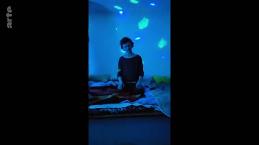 Une personne est assise sur un lit dans une pièce sombre, baignée par des projections de lumières bleues, évoquant une atmosphère introspective ou artistique.
