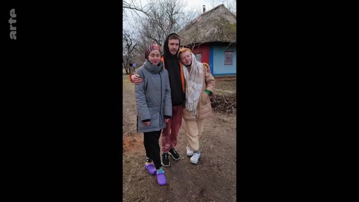 Trois jeunes personnes, deux femmes et un homme, posent pour une photo devant une maison traditionnelle ukrainienne au toit de chaume, dans un contexte de conflit en Ukraine.