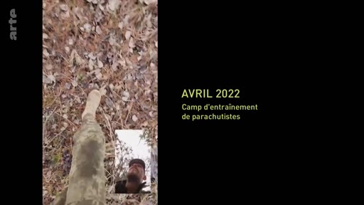 Un soldat en tenue de camouflage marche dans une zone boisée lors d'un camp d'entraînement de parachutistes en avril 2022. L'image montre une vue subjective des jambes du soldat avec une incrustation vidéo montrant son visage.