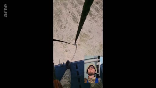 Un soldat ukrainien équipé d'un casque et d'un uniforme de camouflage filme son saut en parachute en vue subjective depuis un avion militaire. L'image montre le sol en contrebas et une incrustation vidéo du visage du soldat.