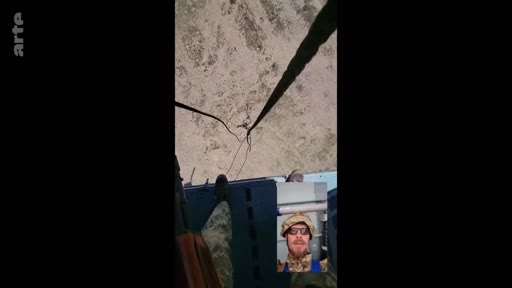 Un soldat en tenue de camouflage et casque tactique filme une vidéo en vue subjective depuis un hélicoptère en vol, montrant le sol en contrebas. Cette séquence illustre le quotidien des militaires en opération ou en entraînement.