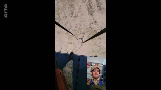 Un soldat en tenue de camouflage filme une vue aérienne depuis un hélicoptère, avec une incrustation vidéo montrant son visage en gros plan. Cette séquence illustre le quotidien et les déplacements des militaires en opération.