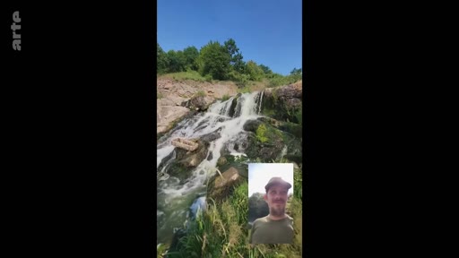 Un homme barbu avec une casquette participe à un appel vidéo en extérieur, avec une cascade en arrière-plan, illustrant un moment de loisir et de connexion à distance.