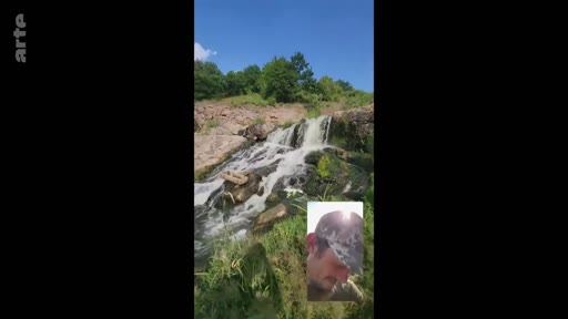 Un soldat en uniforme et casquette, filmé en gros plan via un appel vidéo, montre une cascade naturelle en arrière-plan. Cette séquence illustre le quotidien ou les moments de pause d'un militaire sur le terrain.
