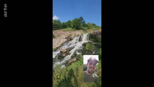 Un homme souriant participe à un appel vidéo en extérieur, avec une cascade en arrière-plan, illustrant un moment de loisir et de connexion à distance.