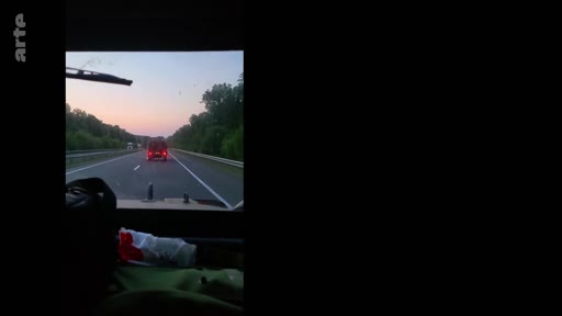 Vue subjective depuis l'intérieur d'un véhicule circulant sur une route bordée d'arbres au crépuscule, illustrant un déplacement dans un contexte de conflit ou de zone de tension.