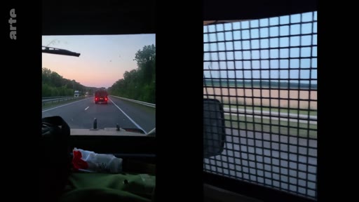 Vue subjective depuis l'intérieur d'un véhicule blindé ou sécurisé, avec une grille de protection sur la fenêtre latérale, circulant sur une route en zone de conflit ou en zone sécurisée.