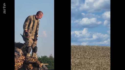 Un soldat ukrainien portant un uniforme de camouflage, des genouillères et des gants tactiques est debout sur un véhicule blindé dans un paysage rural. Cette image illustre la présence militaire sur le terrain dans le contexte du conflit en Ukraine.