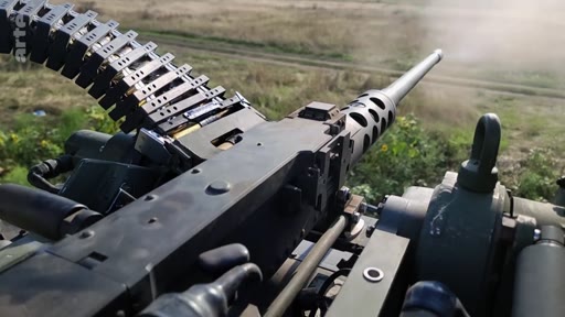 Vue à la première personne d'une mitrailleuse lourde Browning M2 en train de tirer, illustrant des opérations militaires sur le terrain. L'image montre le mécanisme de l'arme et la bande de munitions en mouvement lors d'un engagement armé.