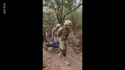 Des soldats ukrainiens en tenue de combat évacuent un camarade blessé sur une civière à travers une zone boisée, illustrant les opérations de secours médical sur le front en Ukraine.