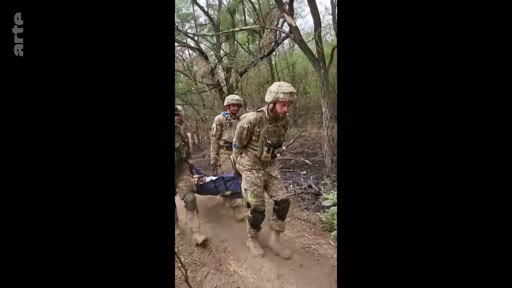 Des soldats ukrainiens en tenue de camouflage et casques militaires transportent un camarade blessé sur une civière à travers une zone boisée, illustrant les opérations de secours médical sur le front en Ukraine.