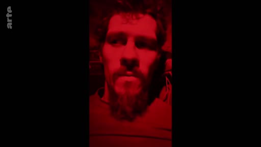 Un homme barbu est filmé en gros plan sous un éclairage rouge saturé, dans une séquence du documentaire 'Reflections on the front line'.