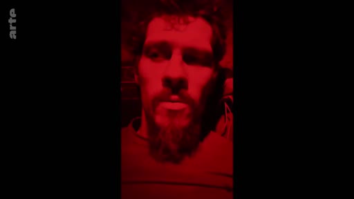 Gros plan sur un homme barbu filmé dans une ambiance sombre avec un éclairage rouge intense, évoquant une situation de tension ou de conflit sur le front.