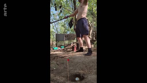 Un homme torse nu pratique le tir à l'arc en extérieur dans un environnement boisé, illustrant une scène de vie quotidienne ou d'entraînement dans le cadre du documentaire 'Comrades, war, and the search for normality'.