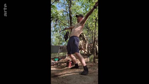 Un homme torse nu, portant une casquette, effectue des exercices d'étirement en plein air dans une zone boisée, dans le cadre d'un reportage sur la vie quotidienne en temps de guerre.