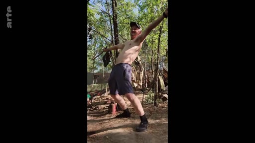 Un homme torse nu, portant une casquette et des shorts, prend une pose dynamique dans un environnement forestier, illustrant une recherche de normalité dans un contexte de guerre.