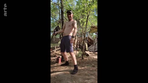 Un homme torse nu, portant une casquette et un short, se tient debout dans un campement improvisé en forêt, illustrant le quotidien des combattants dans le cadre du documentaire Comrades, war, and the search for normality.