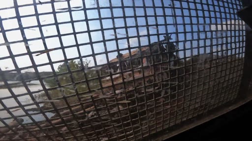 Vue à travers la grille de protection d'un véhicule militaire circulant dans un village ukrainien libéré, montrant des maisons endommagées et un paysage rural dévasté par le conflit.