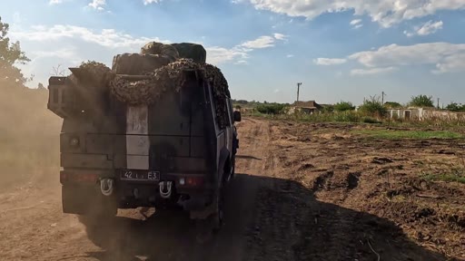 Un véhicule militaire tout-terrain, chargé de matériel et recouvert d'un filet de camouflage, roule sur une piste en terre dans un village libéré. La scène illustre les opérations de sécurisation et de logistique dans les zones récemment reprises lors du conflit en Ukraine.