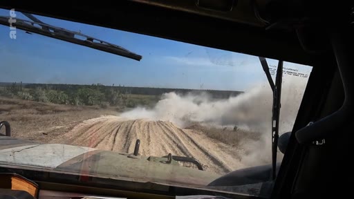 Vue subjective depuis le poste de pilotage d'un véhicule militaire en mouvement sur une piste en terre, soulevant un important nuage de poussière dans un paysage dégagé.