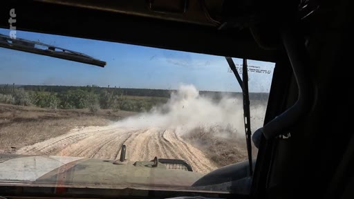 Vue subjective depuis le cockpit d'un véhicule militaire en mouvement sur une piste en terre, soulevant un nuage de poussière dans un paysage rural. Cette image illustre le quotidien des soldats sur le terrain lors d'opérations militaires.