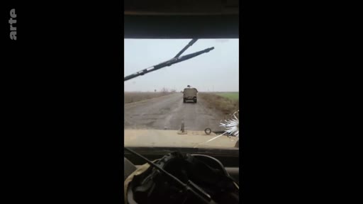 Vue depuis l'intérieur d'un véhicule militaire suivant un autre camion sur une route rurale, illustrant le quotidien des soldats sur le terrain.