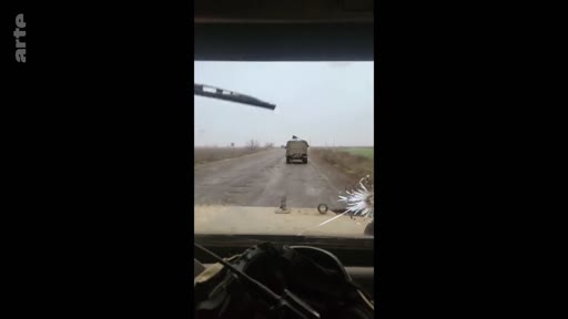 Vue subjective depuis l'intérieur d'un véhicule blindé ou militaire circulant sur une route de campagne, montrant un autre véhicule militaire devant. Cette image illustre le quotidien des soldats sur le terrain dans un contexte de conflit.