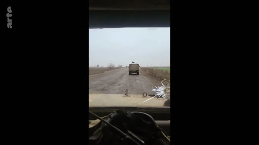 Vue depuis l'intérieur d'un véhicule suivant un autre véhicule militaire sur une route rurale, illustrant le contexte de conflit ou de patrouille.