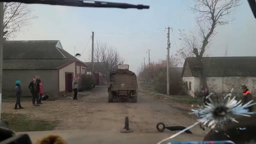 Un véhicule militaire circule sur une route de terre dans un village ukrainien, observé depuis l'intérieur d'un autre véhicule avec un impact de balle sur le pare-brise. Des civils sont visibles sur le bas-côté de la route alors que le convoi progresse dans une zone rurale.