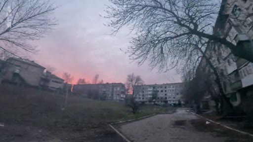 Vue d'ensemble d'un quartier résidentiel avec des immeubles d'habitation dégradés, probablement dans une zone de conflit, sous un ciel crépusculaire.