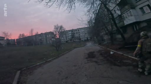 Un soldat ukrainien portant un équipement militaire et un brassard jaune patrouille dans une zone résidentielle endommagée par les combats, sous un ciel crépusculaire. La scène montre l'impact de la guerre sur les infrastructures urbaines en Ukraine.