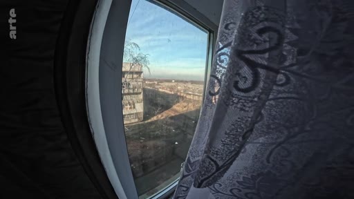 Vue en grand angle depuis l'intérieur d'un appartement montrant un paysage urbain marqué par les destructions de la guerre, avec un immeuble endommagé visible à travers la fenêtre. Cette image illustre le quotidien des civils vivant sur la ligne de front.