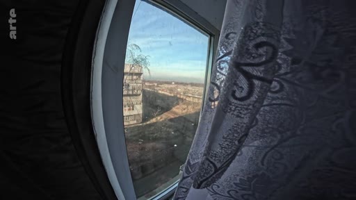 Vue en grand angle depuis une fenêtre montrant un paysage urbain marqué par les stigmates de la guerre, avec des immeubles dégradés dans un quartier résidentiel.