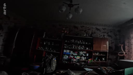 Vue intérieure d'une pièce sombre et encombrée, typique d'un logement dans une zone de conflit, illustrant les conditions de vie sur la ligne de front. On distingue des étagères remplies d'objets personnels et un lustre au plafond.
