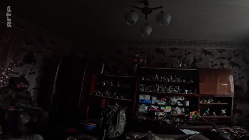 Un soldat en tenue de camouflage est assis dans une pièce sombre et encombrée, illustrant la vie quotidienne sur la ligne de front. L'ambiance est austère et montre les conditions de vie précaires des militaires en zone de conflit.