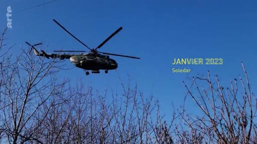 Un hélicoptère militaire survole des arbres dénudés à Soledar en janvier 2023, illustrant le conflit en Ukraine. L'image porte une mention textuelle précisant la date et le lieu de l'événement.