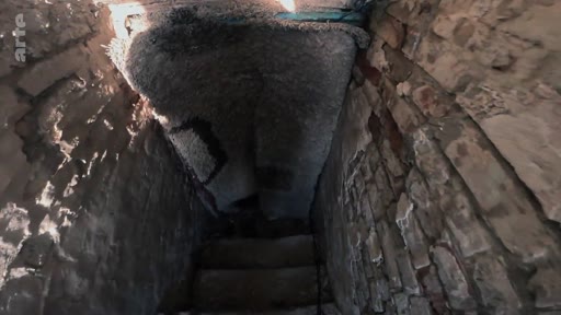 Vue en contre-plongée d'un escalier en pierre menant vers une cave souterraine sombre, illustrant les conditions de vie précaires d'un soldat dans un abri de fortune.