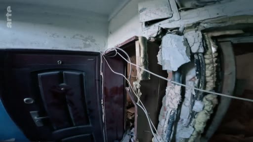 Vue intérieure d'un appartement ou d'un couloir avec une porte d'entrée sombre et des murs dégradés, montrant les stigmates de destructions liées à un conflit armé. La scène illustre les dommages structurels subis par les habitations lors d'opérations militaires.