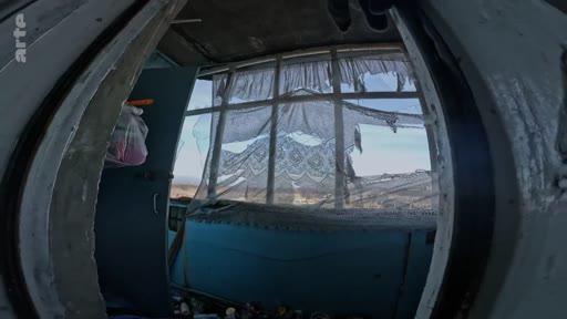 Vue intérieure d'une pièce en ruine avec des fenêtres brisées et des rideaux en lambeaux, témoignant des destructions causées par un conflit ou une attaque.