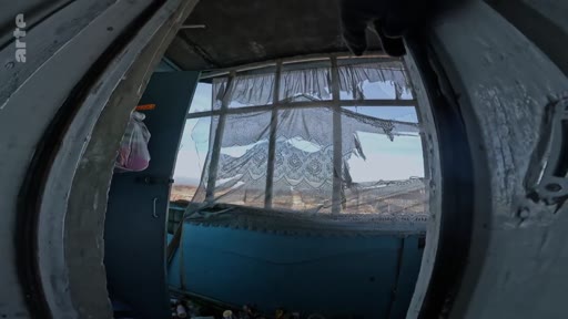 Vue en contre-plongée à travers une fenêtre aux vitres brisées et aux rideaux déchirés dans un bâtiment délabré, évoquant un contexte de conflit ou d'abandon.