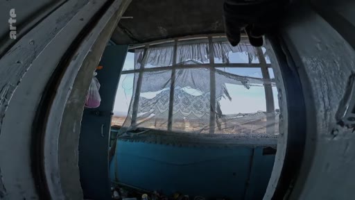 Vue en contre-plongée à travers une fenêtre aux vitres brisées et aux rideaux déchirés, montrant un paysage extérieur dévasté. Cette image illustre les conséquences des combats et des destructions dans une zone de conflit.